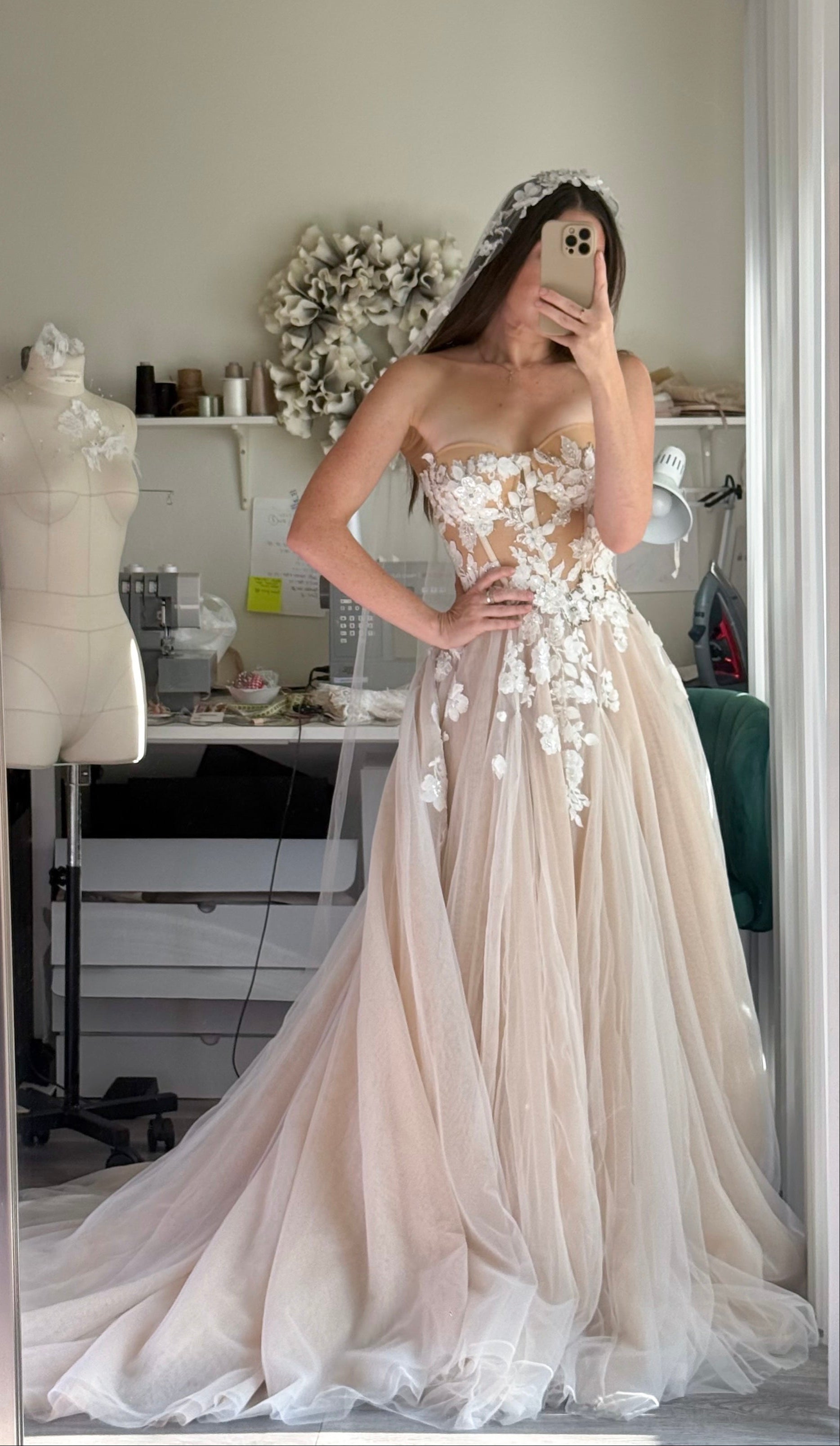 A woman takes a mirror selfie wearing a strapless blush-beige tulle ball gown with 3D floral appliqués on the bodice and cascading down the skirt. The dress features a sweetheart neckline, a fitted bodice, and a full flowing skirt with a train.